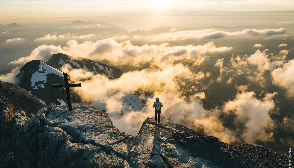 Eine Person steht mit dem Rücken zur Drohnenkamera neben einem Gipfelkreuz. Unterhalb des felsigen Gipfels sind ein paar lockere Wolken zu sehen, aus denen weitere Gipfel mit Schneefeldern durchstechen. | © Black Forest Collective, Quelle: EOFT Media Hub