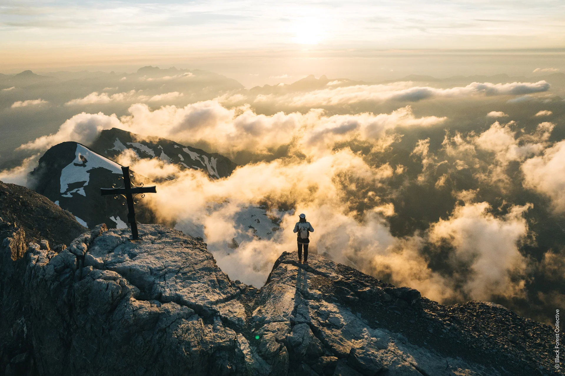 Eine Person steht mit dem Rücken zur Drohnenkamera neben einem Gipfelkreuz. Unterhalb des felsigen Gipfels sind ein paar lockere Wolken zu sehen, aus denen weitere Gipfel mit Schneefeldern durchstechen. | © Black Forest Collective, Quelle: EOFT Media Hub