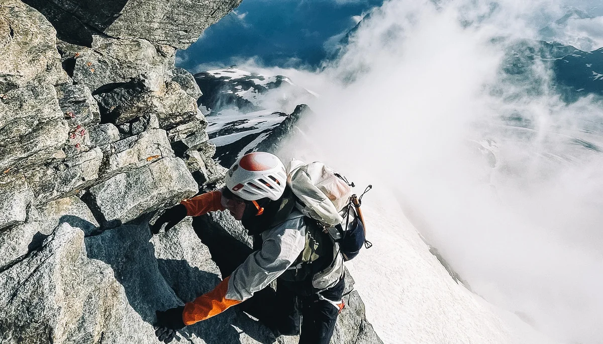 Eine Person klettert einen Quergang im Nachstieg an einer steilen Felswand. Im Hintergrund sieht man eine Schneeflanke, die zum Großteil von Wolken überdeckt ist. | © Black Forest Collective, Quelle: EOFT Media Hub
