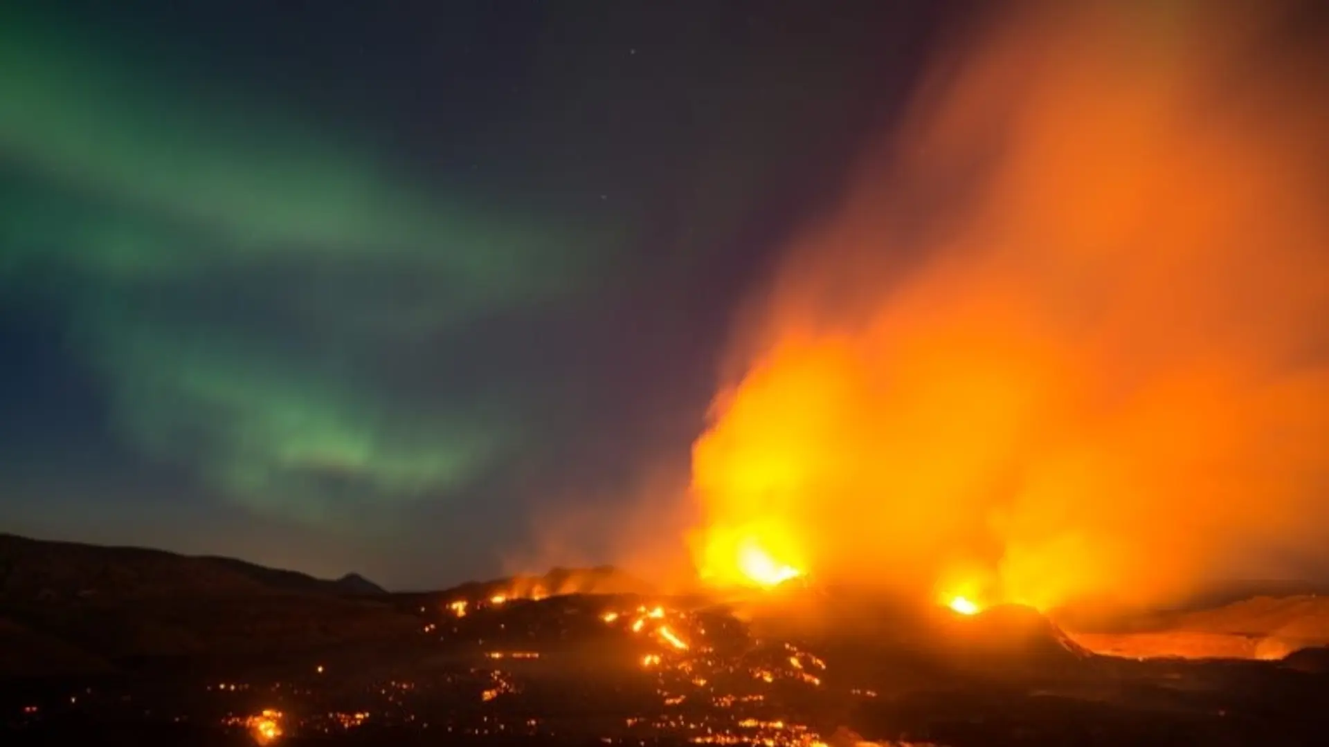 Grün leuchtende Polarlichter am Nachthimmel über einer isländischen Vulkanlandschaft, während glühende Lava und orangfarbene Rauchwolken aus einem aktiven Vulkanausbruch aufsteigen. | © Stephan Schulz