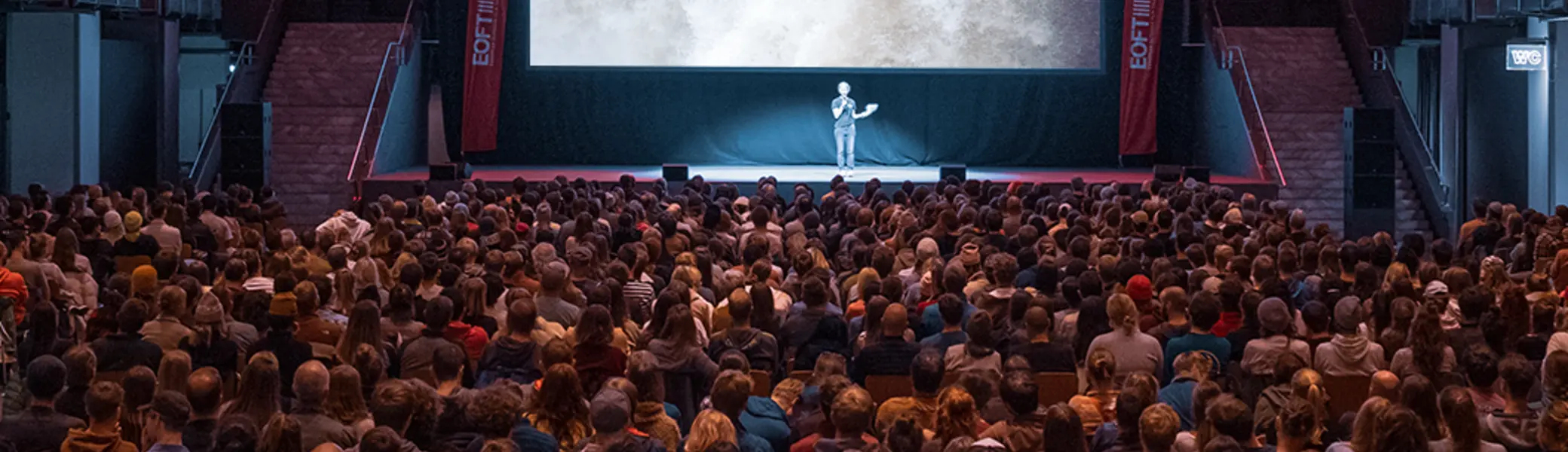 Das Bild zeigt einen dunklen Saal mit einer Leinwand. Davor sitzt eine große Audienz. | © Jana Weinert