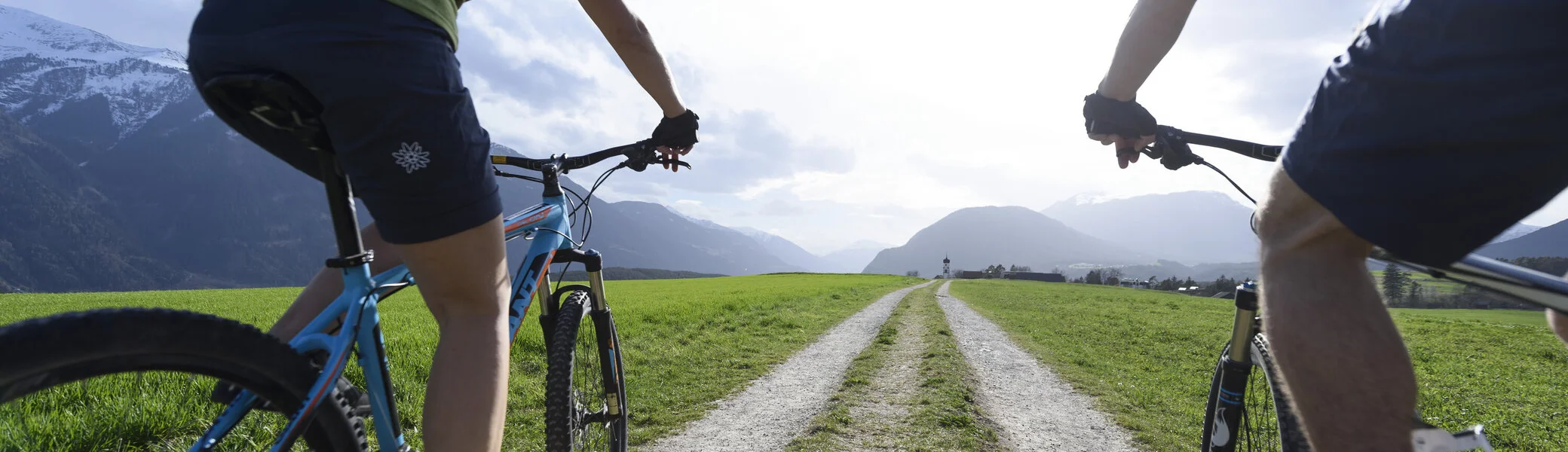Zwei Mountainbiker fahren auf einem Schotterpfade entlang einer grünen Wiese | © DAV/Wolfgang Ehn